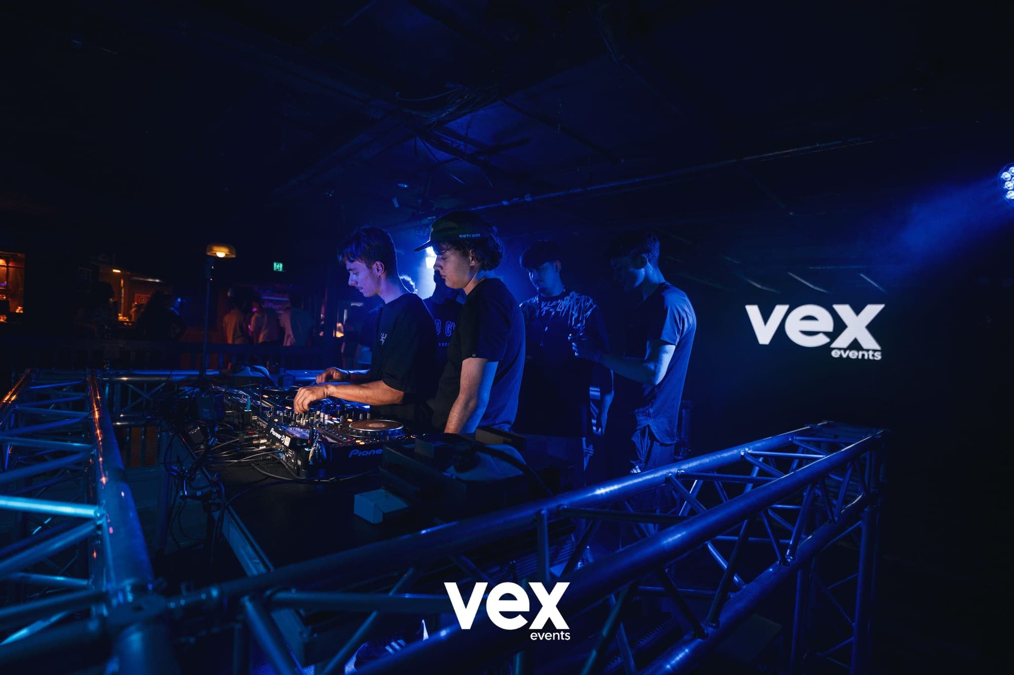 A wide view of DJs, decks and the surrounding room washed in blue light beside a Vex screen