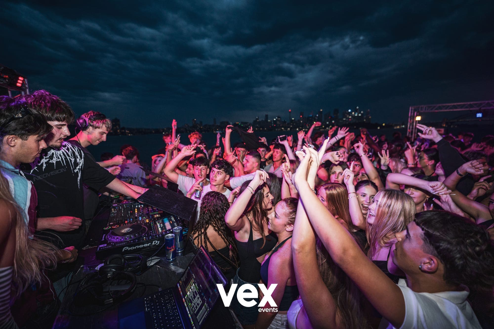 Hands raised across a purple-lit boat dancefloor beneath a deep blue, cloudy evening sky