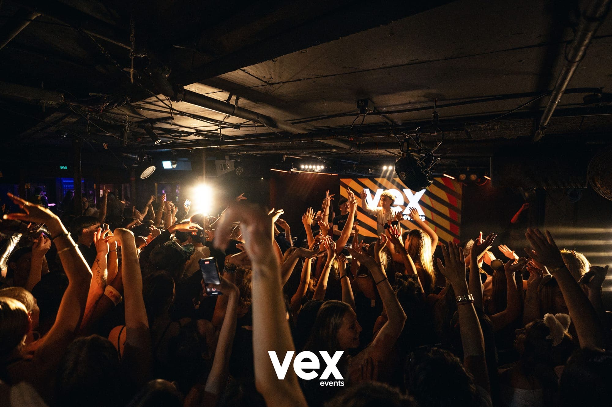 A packed indoor crowd with hands raised towards warm stage lights and a Vex screen