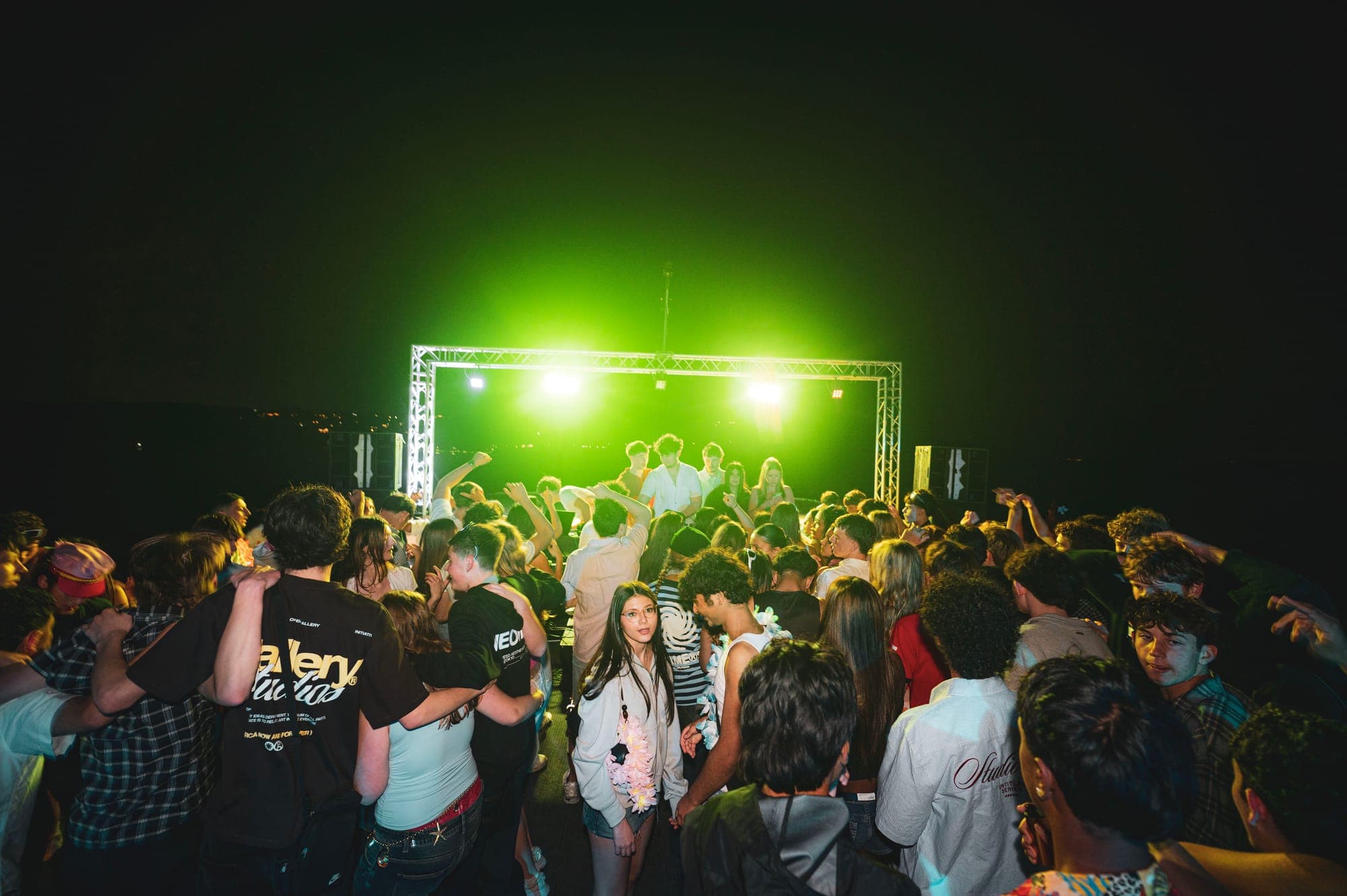 An outdoor crowd facing a DJ platform and rectangular lighting truss glowing green against the night sky