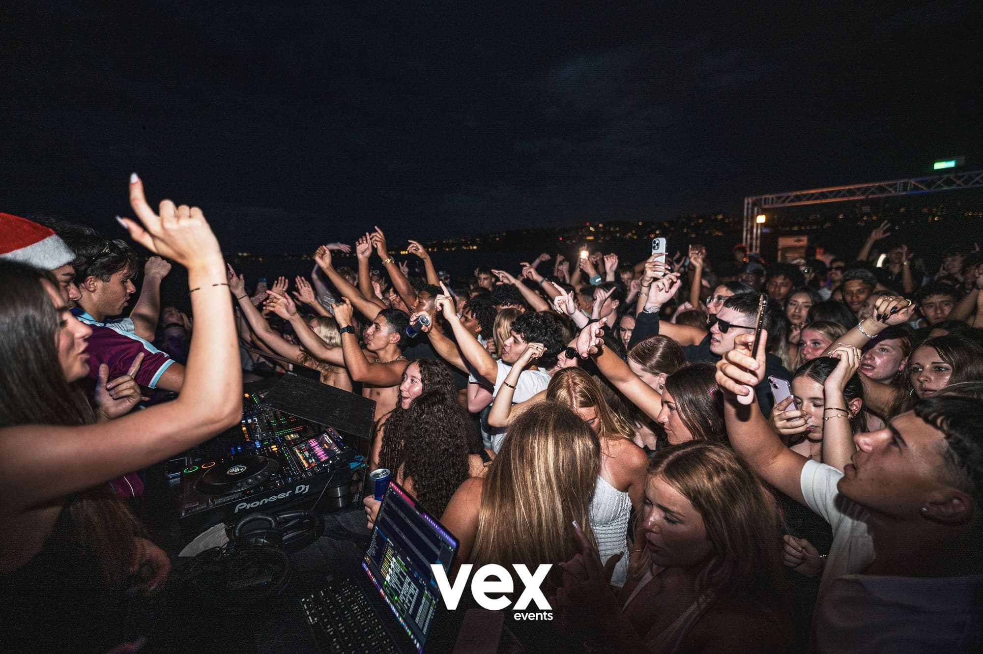 The view across a DJ booth and its mixer towards a full crowd with their hands in the air