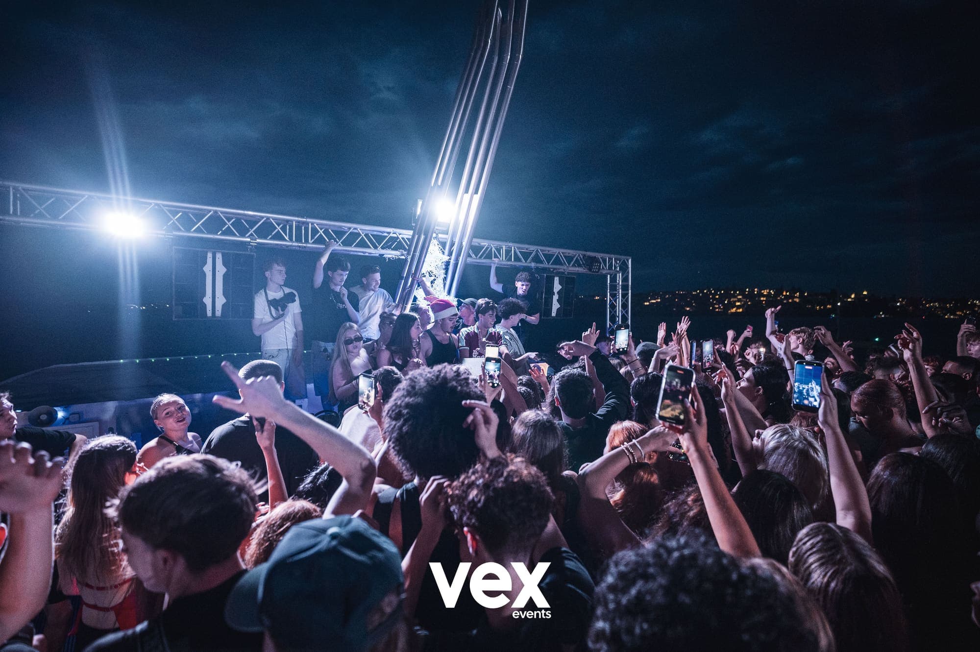A packed outdoor dancefloor at night under a lighting truss, with Sydney lights across the water behind it