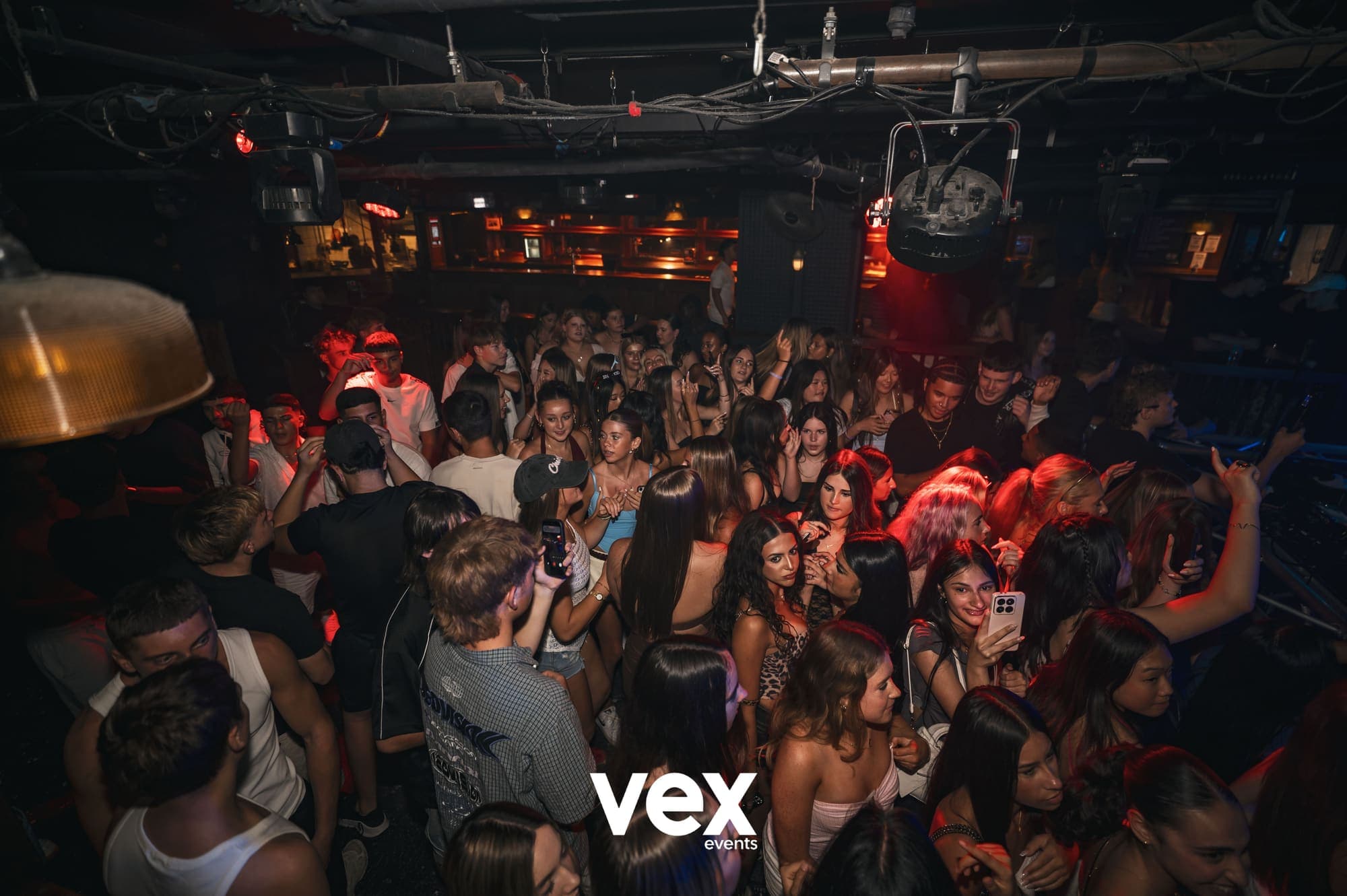 A busy indoor dancefloor lit in deep red, shot from above the crowd