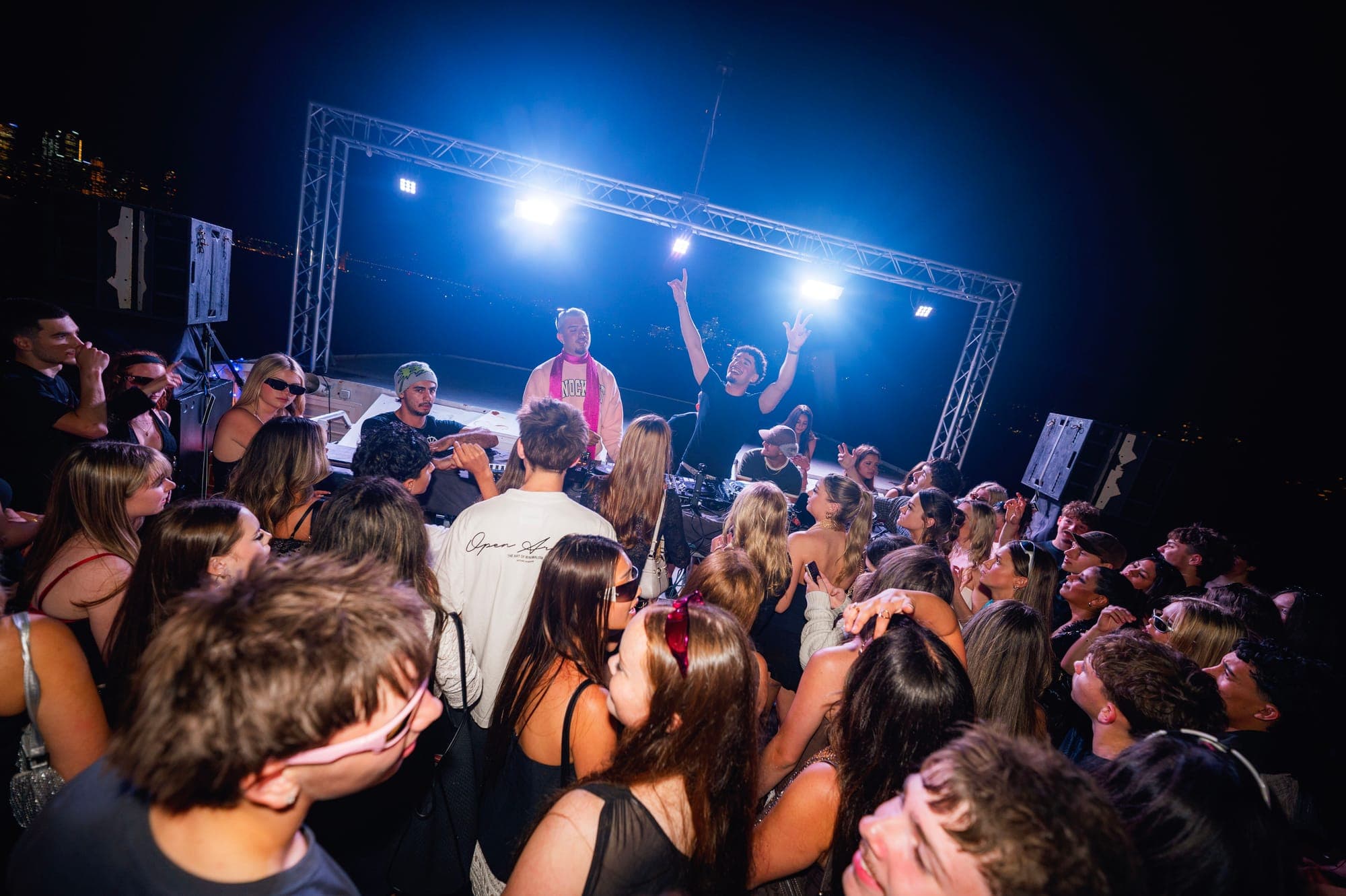 DJs under a blue-lit truss on a boat deck, with a crowd gathered around the decks