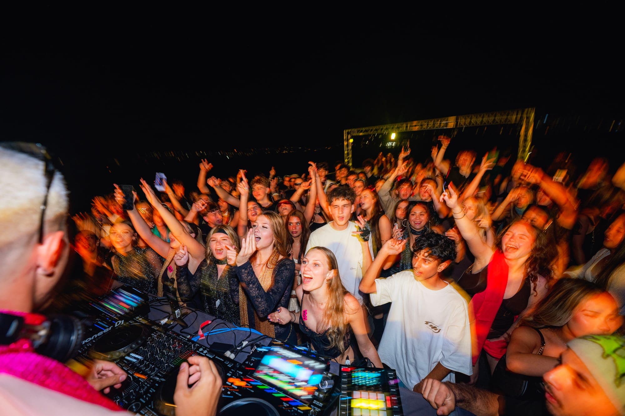 A crowd cheering with hands up in front of the DJ decks on an open boat deck at night