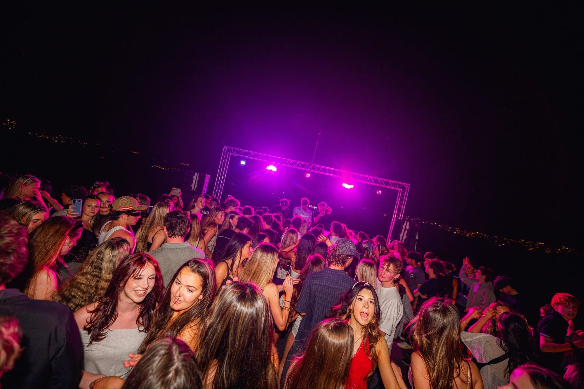 A packed open deck dancing towards a pink-lit DJ stage, with city lights on the water behind